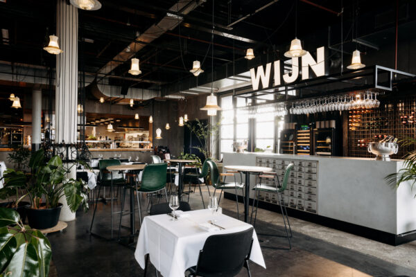 Modern restaurantinterieur aan de Johan Huizingalaan met open keuken, hoge tafels en een opvallende wijnbar met glazenrek en het woord “WIJN” in grote letters.